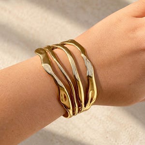 May include: A gold-toned cuff bracelet featuring three distinct, interconnected bands with a smooth, polished, and irregular wavy surface texture that wraps around the wrist, creating a layered and fluid appearance.