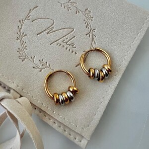 May include: A pair of gold-toned hoop earrings featuring alternating small gold and silver-toned beads threaded onto each hoop. The earrings are displayed on a beige fabric pouch with the text Mila Jewelry printed in a script font with a leaf wreath.