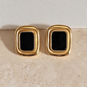 May include: A pair of rectangular earrings featuring a polished gold-tone metal frame with rounded corners and a smooth, solid black rectangular inlay centered within each piece. The design is symmetrical, showcasing a reflective metallic finish.