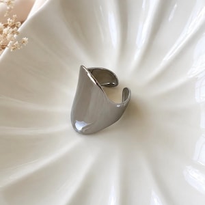 May include: A polished silver-tone metal ring featuring a wide, smooth, and curved open-band design. The ring has a reflective, high-shine finish with a tapered shape that wraps around the finger, showcasing a clean and solid metallic surface.