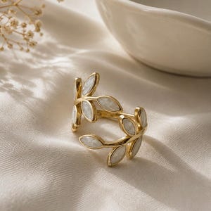 May include: A gold-toned ring featuring a wrap-around design shaped like a leafy vine. The ring is adorned with multiple marquise-shaped white inlays that resemble mother-of-pearl, set within the polished gold metal frame of the leaves.