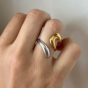 May include: A two-tone open ring featuring a smooth, polished silver-colored band on one side and a curved, gold-colored band on the other. The ring has a fluid, asymmetrical design with rounded edges, creating a continuous, open-ended shape.