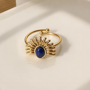 May include: A gold-colored ring with a sunburst design and a deep blue oval stone. The ring is set on a white surface, showcasing its intricate details and elegant style. The ring's adjustable band adds to its versatility.