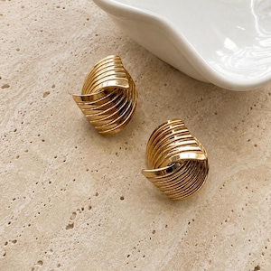 May include: A pair of gold-toned earrings featuring a rounded, sculptural design with multiple parallel, raised ridges that curve inward toward a central point, creating a layered, fan-like appearance with a polished, reflective metallic finish.