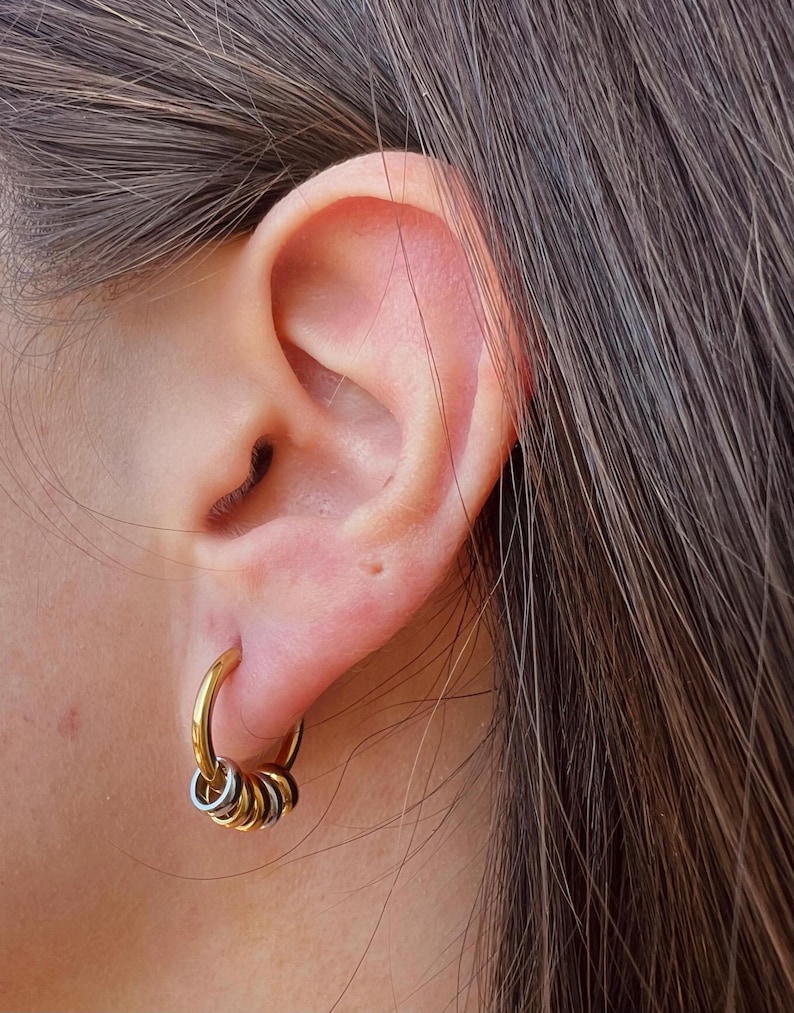 May include: A gold-toned hoop earring featuring a series of smaller, alternating gold and silver-toned rings suspended from the main hoop. The earring is worn in the earlobe, showcasing a multi-layered, metallic design with a polished finish.