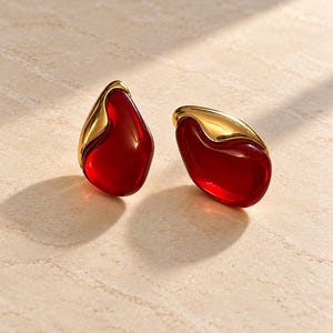 May include: A pair of stud earrings featuring smooth, translucent red, teardrop-shaped stones partially encased in polished gold-tone metal. The earrings have an organic, asymmetrical form with a glossy finish that reflects light.