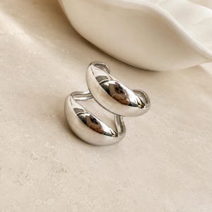 May include: A silver-toned ring featuring a bypass design with two smooth, rounded, bulbous sections that overlap. The metal has a highly reflective, polished finish that captures light and creates distinct highlights across its curved surface.