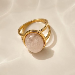 May include: A gold-toned ring featuring a smooth, oval-shaped pale pink gemstone set within a polished metal bezel. The band of the ring is split into two distinct sections that meet at the setting, creating an open, layered appearance.