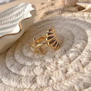 May include: A gold-toned open ring featuring a textured starfish on one end and a ribbed seashell design on the other. The metal has a polished finish, showcasing detailed ridges on the shell and a raised, dotted pattern on the starfish arms.