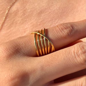 May include: A gold ring featuring a multi-band design with five parallel vertical bands that are crossed by a single diagonal band. The metal has a polished, reflective finish that catches the light, creating a bright and metallic appearance.
