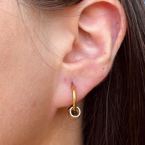 May include: A gold-toned hoop earring featuring a small, dangling circular charm. The earring is secured through the earlobe, showcasing a polished metallic finish and a simple, minimalist design that hangs vertically from the main hoop component.