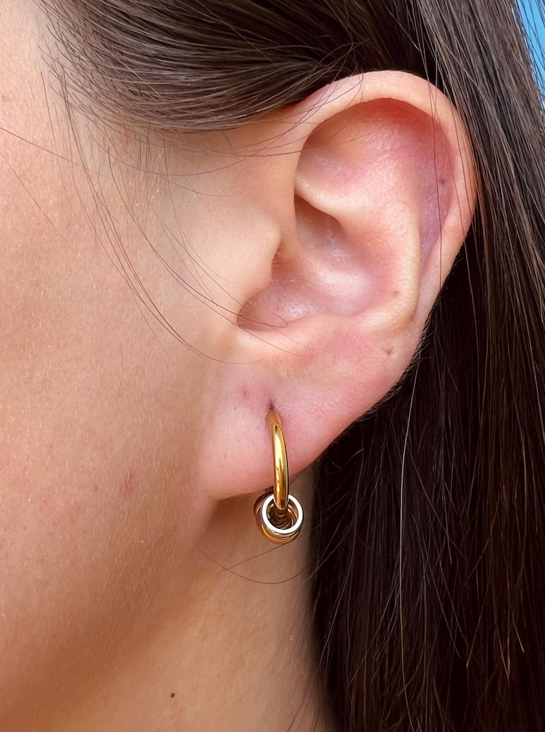 May include: A gold-toned hoop earring featuring a small, dangling circular charm. The earring is secured through the earlobe, showcasing a polished metallic finish and a simple, minimalist design that hangs vertically from the main hoop component.