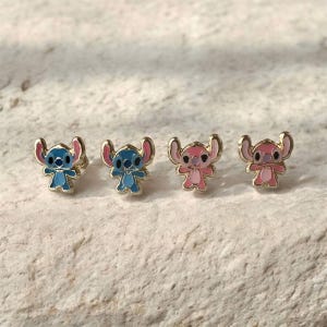 May include: Four small stud earrings featuring the character Stitch from the animated film. Two are blue with gold trim, and two are pink with gold trim. The earrings are displayed on a textured, light-colored surface.