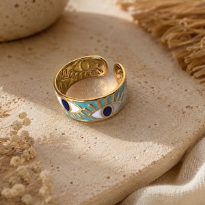 May include: Gold-toned adjustable ring with an eye design. The ring features a light blue enamel band with white and navy blue eye details. The inner band has an etched eye pattern.