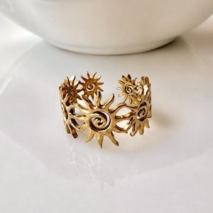 May include: A gold-toned metal ring featuring a repeating openwork pattern of stylized suns. Each sun motif has a central spiral design with radiating pointed rays. The band is open at the back, allowing for adjustment in size and fit.