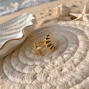 May include: A gold-colored ring with a starfish and seashell design. The ring is on a woven, circular coaster. A white seashell-shaped dish with a gold trim is in the background, along with a starfish and seashells on a sandy beach.