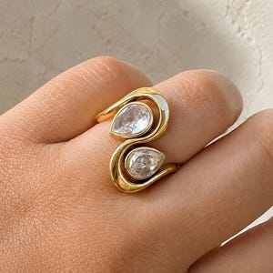 May include: A gold ring featuring a bypass design with two pear-shaped clear stones set in smooth metal frames. The band curves around the finger, creating an open, flowing silhouette that highlights the two faceted, teardrop-shaped gemstones.