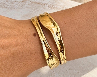 May include: Two gold-toned metal cuff bracelets with a polished, reflective finish and an irregular, organic wavy shape. The bracelets feature a smooth, undulating surface that catches the light, creating a fluid appearance on the wrist.