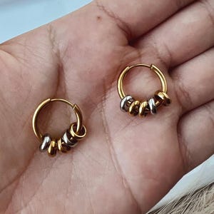 May include: A pair of hoop earrings featuring a gold-toned circular frame. Each hoop is adorned with three small, alternating metallic rings in gold and silver tones that slide along the main hoop structure, creating a layered and textured appearance.