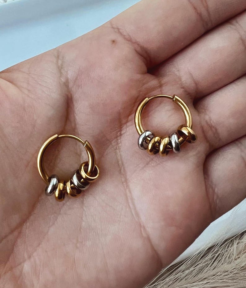 May include: A pair of hoop earrings featuring a gold-toned circular frame. Each hoop is adorned with three small, alternating metallic rings in gold and silver tones that slide along the main hoop structure, creating a layered and textured appearance.