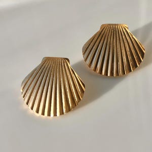 May include: A pair of gold-toned metal earrings shaped like fan-style seashells. Each earring features a series of vertical ridges radiating from a flat top edge to a rounded bottom, creating a textured, metallic surface with a polished finish.
