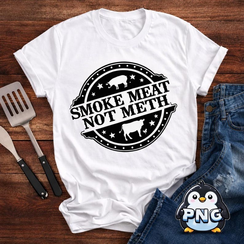 White T-shirt mockup on a rustic wood background featuring a black Smoke Meat Not Meth design with grilling tools, jeans, and the PNGuino penguin logo in the corner