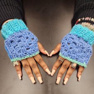 May include: Crocheted fingerless gloves in shades of blue and teal. The gloves have a textured pattern and are trimmed with a light green border. The image shows the gloves on a person's hands, with long, light-colored nails and various rings and bracelets.