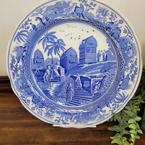 Op de afbeelding: Een blauw-wit decoratief bord met een gedetailleerde landschapsweergave. Het bord toont gebouwen, figuren en dieren in een klassieke, vintage stijl. De rand heeft een geschulpte rand.