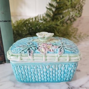 May include: A turquoise ceramic basket-shaped trinket box with a lid. The lid features embossed floral and leaf designs in pink and green. The box has a woven basket texture.