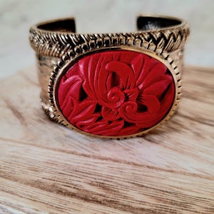May include: A wide, gold-toned cuff bracelet with a central oval of carved red material. The red oval features a floral design. The bracelet has a textured, woven pattern around the edges.