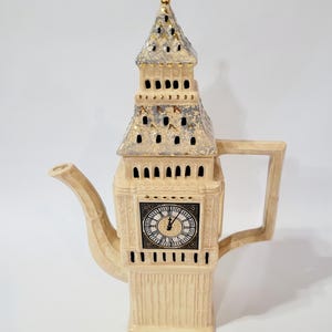 Puede incluir: Una tetera de cerámica de color crema con forma de la Torre Elizabeth, que presenta una esfera de reloj con números romanos negros, un remate dorado en la parte superior y un techo texturizado con detalles metálicos plateados y dorados.