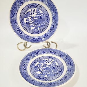 May include: Two round ceramic plates featuring a blue and white willow pattern. The design includes intricate illustrations of pagodas, trees, and birds within a central circular border, surrounded by a decorative geometric and floral rim.