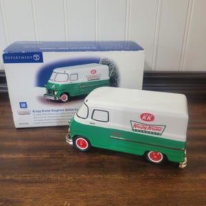 May include: A miniature Krispy Kreme delivery van model featuring a white upper body and a green lower body with red wheels. The side of the van displays the red and green Krispy Kreme logo. It is positioned in front of its original blue and white box.