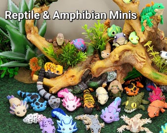 May include: A collection of small, articulated reptile and amphibian figurines in various colors, including axolotls, lizards, and frogs. The text "Reptile & Amphibian Minis" and "Joyful Layers" is visible. The figures are arranged on a green surface.