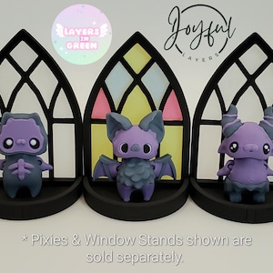 May include: Three whimsical figurines in front of black arched window stands. The figurines are purple and gray, resembling a creature, a bat, and a horned demon. The windows have colorful stained-glass designs. Text on the image says "Layers in Green" and "Joyful Layers".