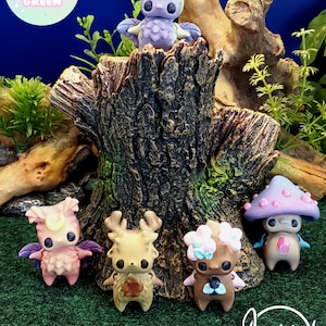 May include: Five handmade miniature figurines, each with a unique design and color scheme. The figurines are displayed on a tree stump with green grass in the background. The figurines include a purple owl, a pink bird, a brown deer, a pink and blue fairy, and a purple mushroom creature.