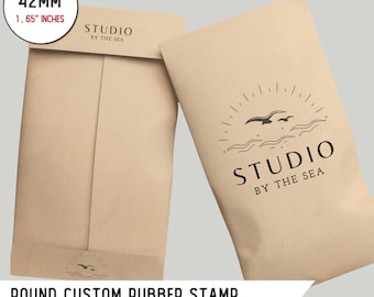Könnte beinhalten: Zwei braune Papiertüten mit einem runden, individuellen Gummistempel. Der Stempel hat einen Durchmesser von 42 mm und zeigt ein Logo mit dem Text "Studio by the Sea" und einer Grafik mit einer Sonne, Wellen und Möwen. Der Text "Runder individueller Gummistempel Senden Sie uns Ihr Logo" ist unter den Tüten gedruckt.
