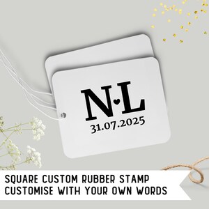 May include: Two white square tags with the initials "N L" and a heart symbol, along with the date "31.07.2025" in black. The tags are connected with white string. The text "SQUARE CUSTOM RUBBER STAMP CUSTOMISE WITH YOUR OWN WORDS" is at the bottom.