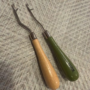 May include: Two latch hook tools with metal shafts and hooks. One tool features a light-colored natural wood handle, while the other has a smooth, olive green painted handle with the black text XU printed near the base of the handle.