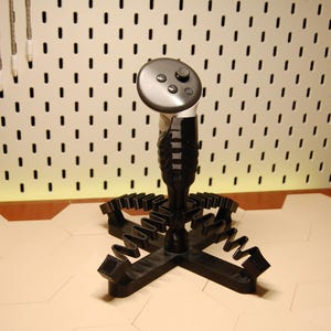 May include: A black 3D-printed controller stand featuring a central vertical post with a textured grip and a four-legged base with notched edges designed to hold a gaming controller. The stand is shown with a gray controller resting on top.