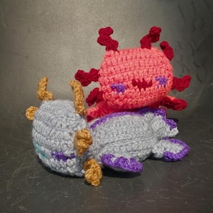 Puede incluir: Dos animales de peluche tejidos a crochet, uno un ajolote rosa con branquias rojas y el otro un dragón gris con detalles morados.