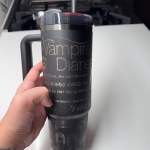 Op de afbeelding: Een zwarte roestvrijstalen beker met een zwarte handgreep en een zwarte deksel. De beker heeft een citaat uit de tv-serie "The Vampire Diaries" gegraveerd op de zijkant. Het citaat luidt: "Wanneer het echt is, kun je niet wachten. Je zult altijd kiezen wat je liefhebt. Bewijs me dat dit voor altijd is. En eerlijk gezegd, toen ik..."