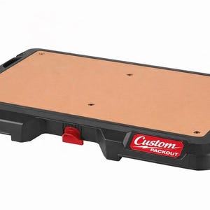 Peut inclure: Base rectangulaire noire et beige avec le texte "Custom PACKOUT" en rouge et blanc. La base a un bouton de déverrouillage rouge et est conçue pour l'organisation des outils. La surface supérieure est beige et comporte plusieurs trous pré-percés.