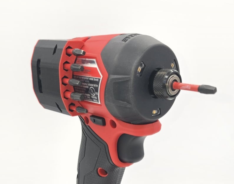 May include: A red and black cordless impact driver with a black grip. The tool has a black head with a red bit inserted. Several bits are stored on the side. The tool is designed for professional use.