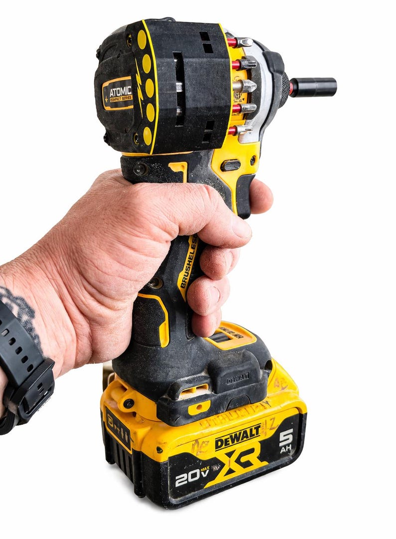 May include: A black and yellow cordless drill with a battery pack. The drill is held in a hand and has the brand name "DEWALT" and "ATOMIC" on it. The drill has a variety of bits attached.
