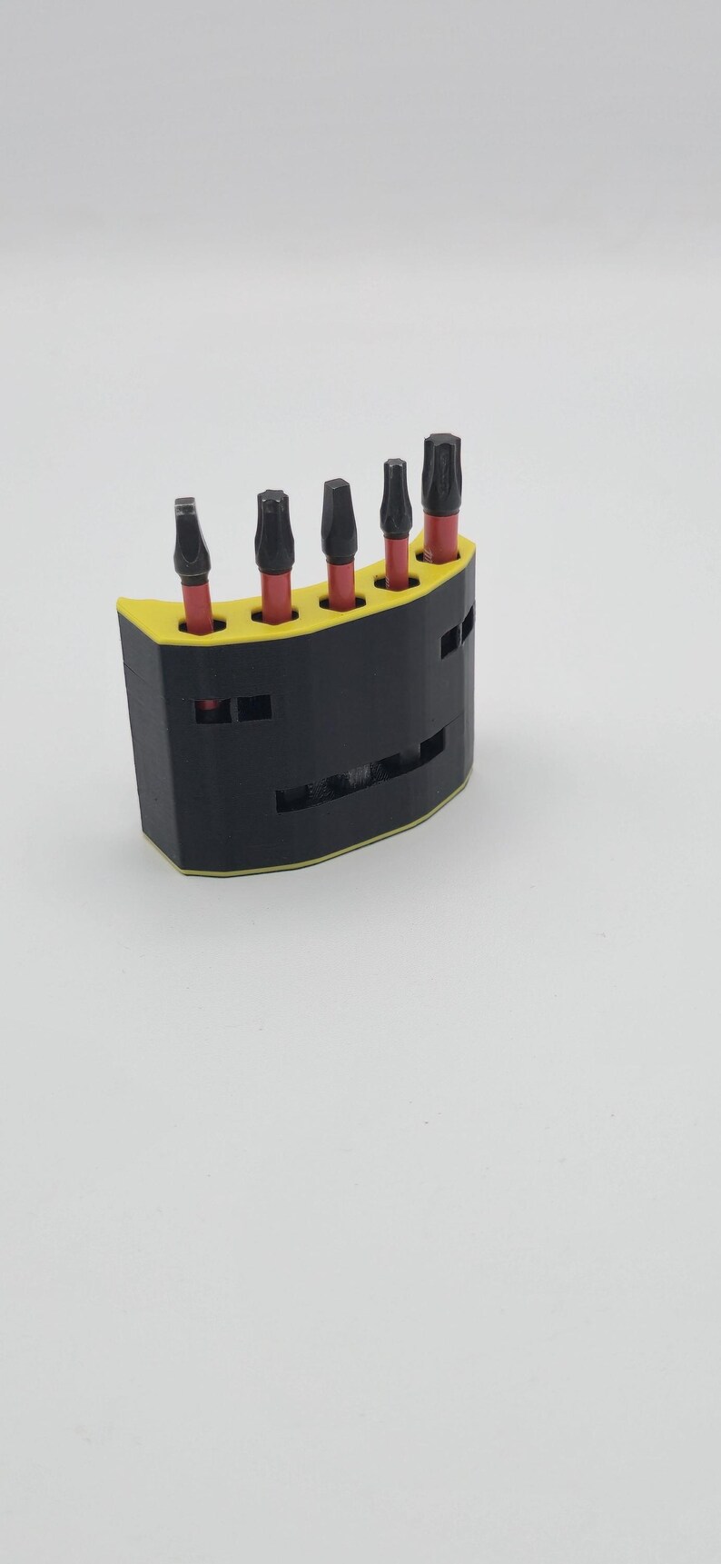 May include: A black and yellow tool holder with five black and red bits. The holder is semi-circular with a yellow top and a black base. The bits are arranged in a row, each with a black head and a red shaft.