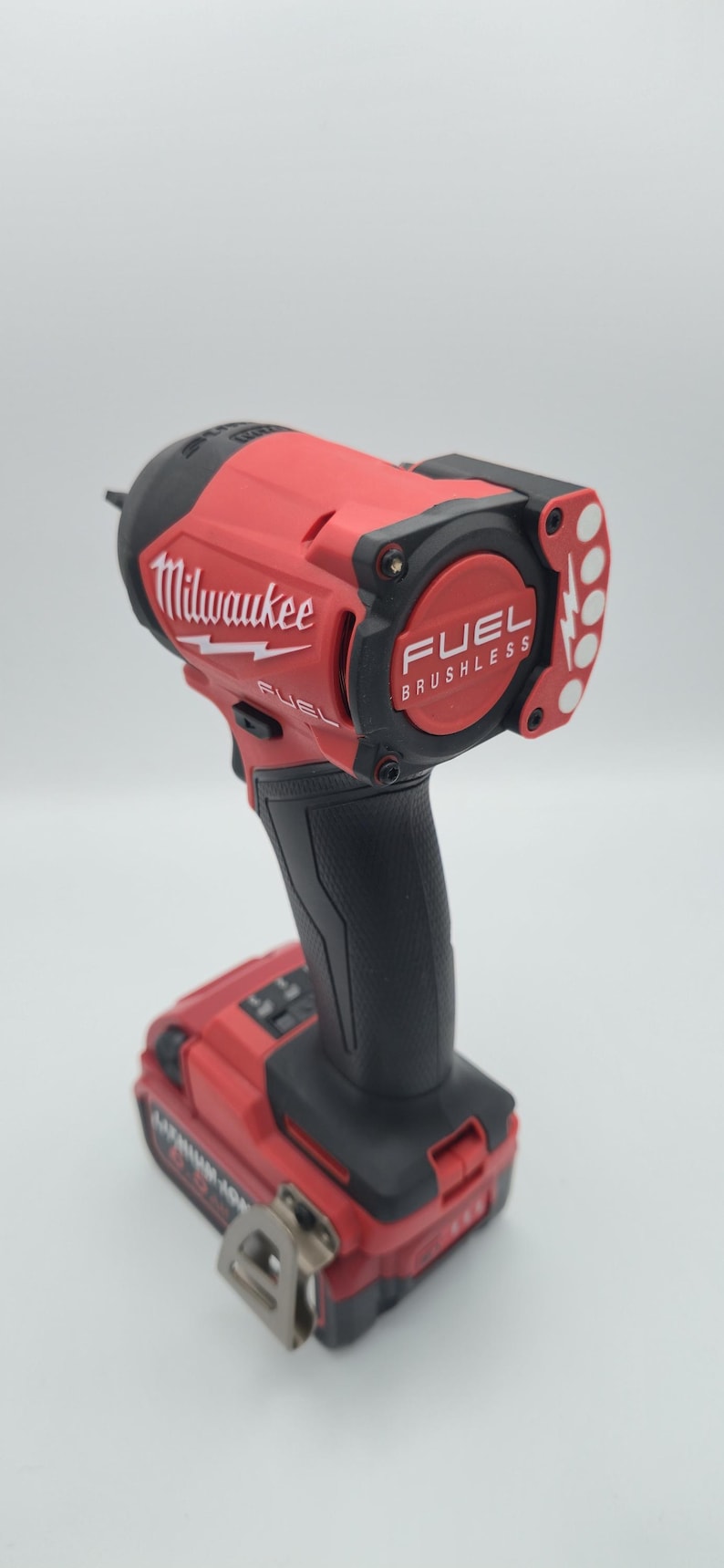 May include: A red and black Milwaukee FUEL brushless impact wrench. The tool features the Milwaukee logo and the text "FUEL BRUSHLESS". The impact wrench has a black handle and a battery pack attached.