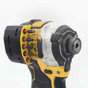 May include: A yellow and black cordless impact driver with a black and yellow bit holder. The tool has a silver head and a black grip. The bit holder contains several red and black bits. The tool has the text "TYPE 1" on it.