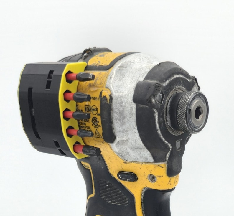 May include: A yellow and black cordless impact driver with a black and yellow bit holder. The tool has a silver head and a black grip. The bit holder contains several red and black bits. The tool has the text "TYPE 1" on it.