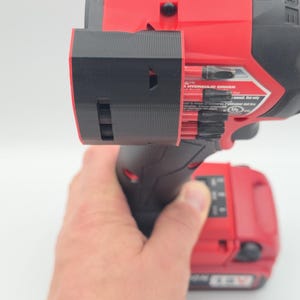 May include: Red and black power drill with a black attachment. The drill has the text "M18 SURGE" and "HYDRAULIC DRIVER" printed on it. The drill is held by a person.